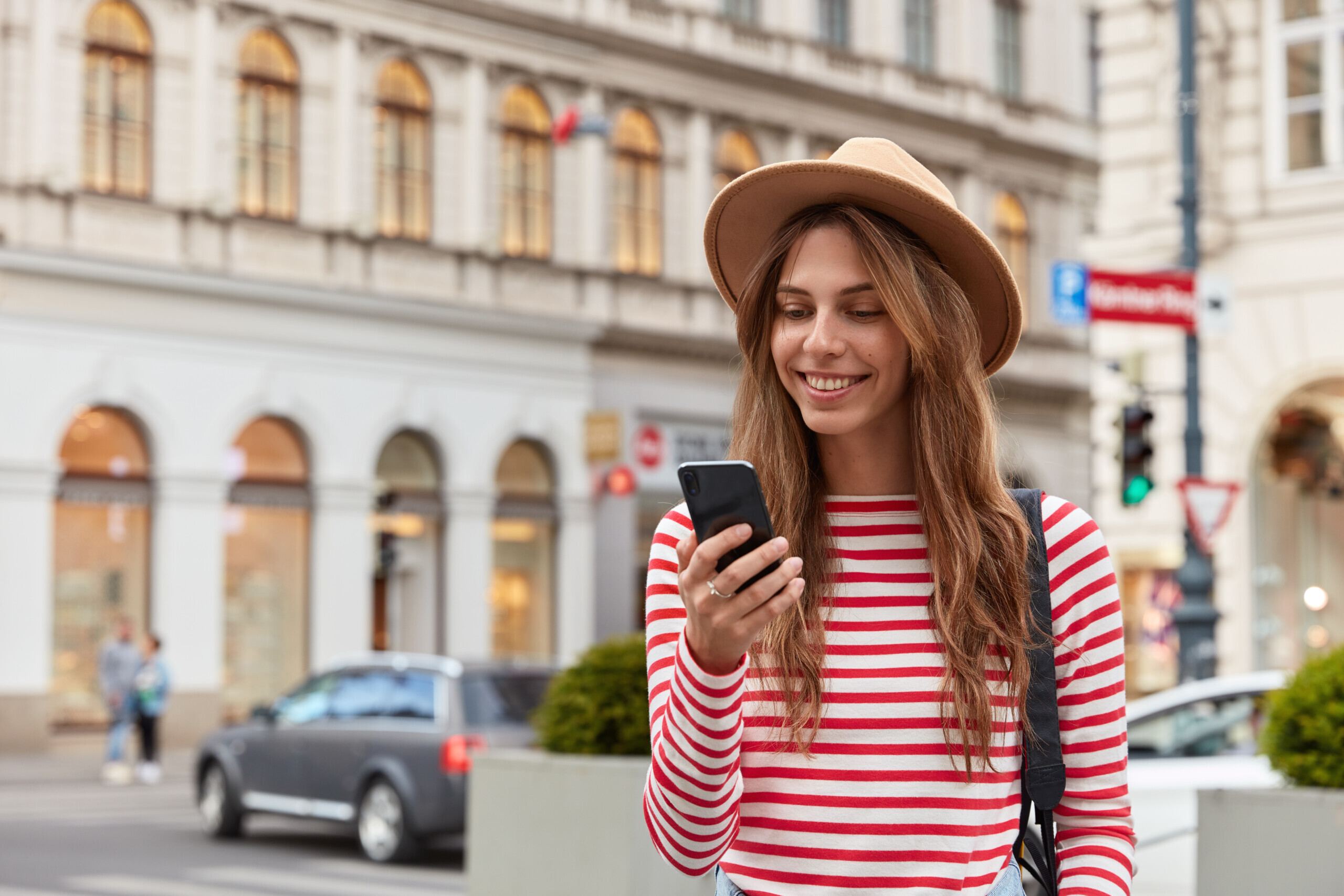 Smiling young European woman enjoys recreation time, watches interesting video on website, wears stylish headgear and striped sweater, has joyful expression, poses over urban setting, chats online