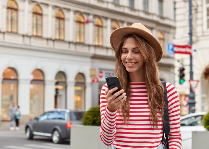 Smiling young European woman enjoys recreation time, watches interesting video on website, wears stylish headgear and striped sweater, has joyful expression, poses over urban setting, chats online